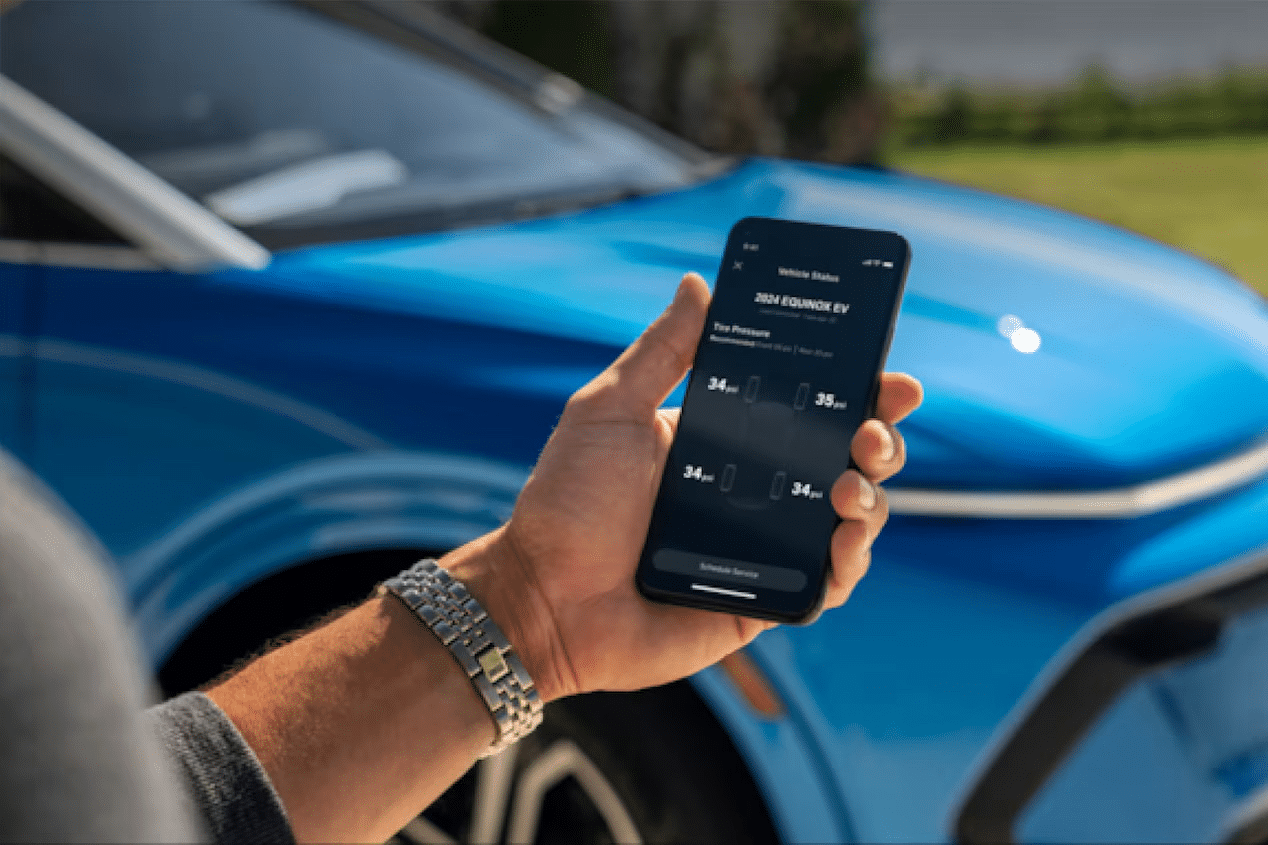 Person holding a smartphone displaying 2024 Equinox EV tire pressure, next to a blue car.