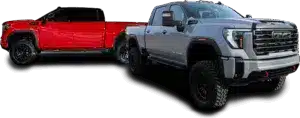 Two pick-up trucks, one red and one gray, are parked on a runway or a large parking lot. The red truck is at an angle that makes it look farther away and smaller, while the gray truck is closer and larger. Behind them, a city skyline with tall buildings can be seen under a blue sky with scattered cloud