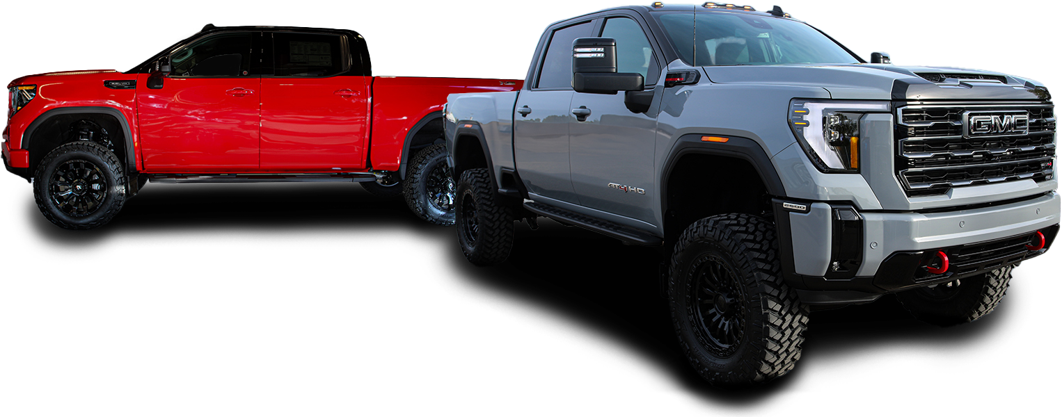 Two pick-up trucks, one red and one gray, are parked on a runway or a large parking lot. The red truck is at an angle that makes it look farther away and smaller, while the gray truck is closer and larger. Behind them, a city skyline with tall buildings can be seen under a blue sky with scattered cloud